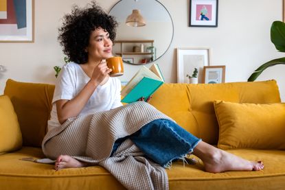 Pensive relaxed African American woman reading a book at home, drinking coffee sitting on the couch
