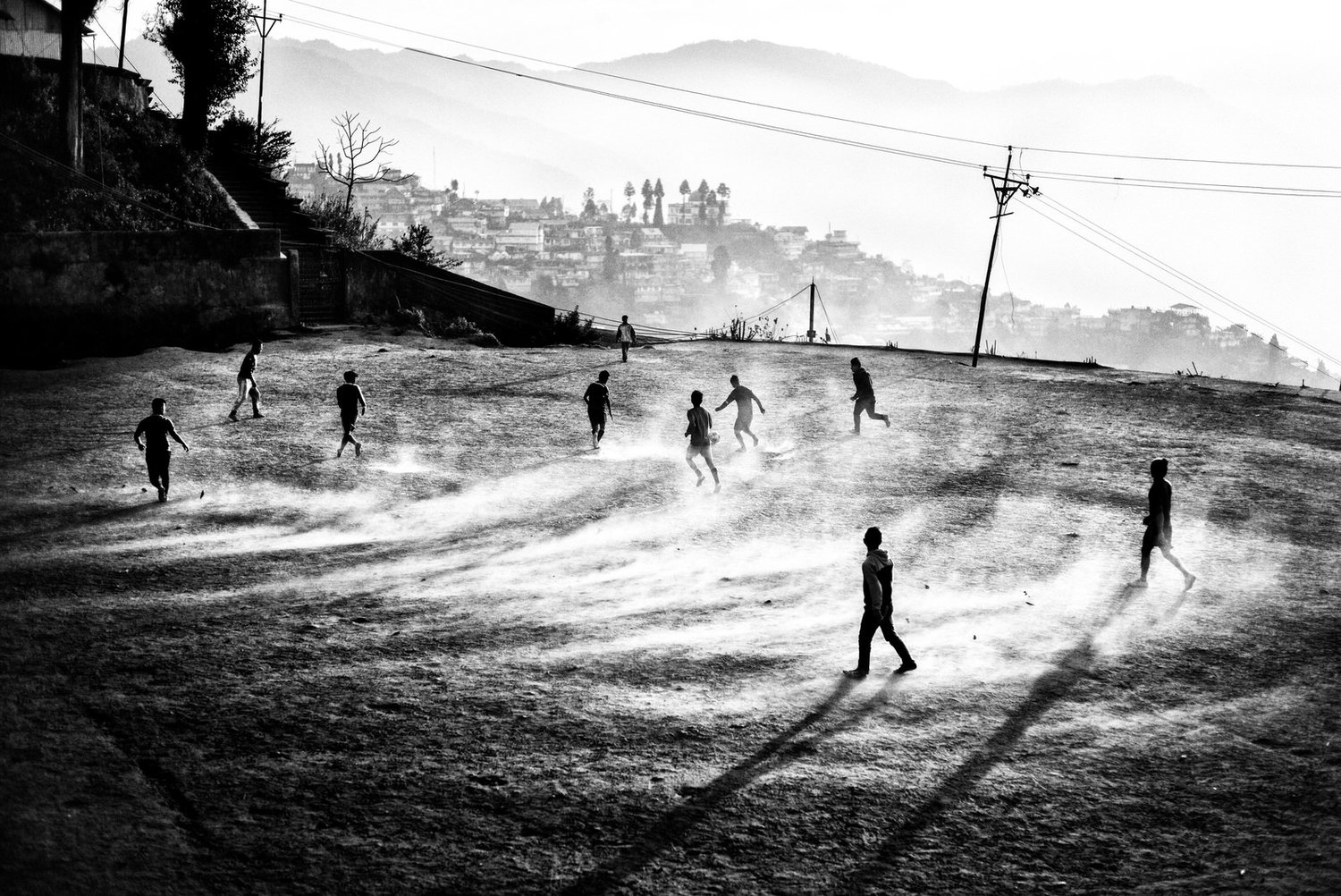 A black-and-white image of several boys playing soccer on a dusty field, with mountains and a village in the background