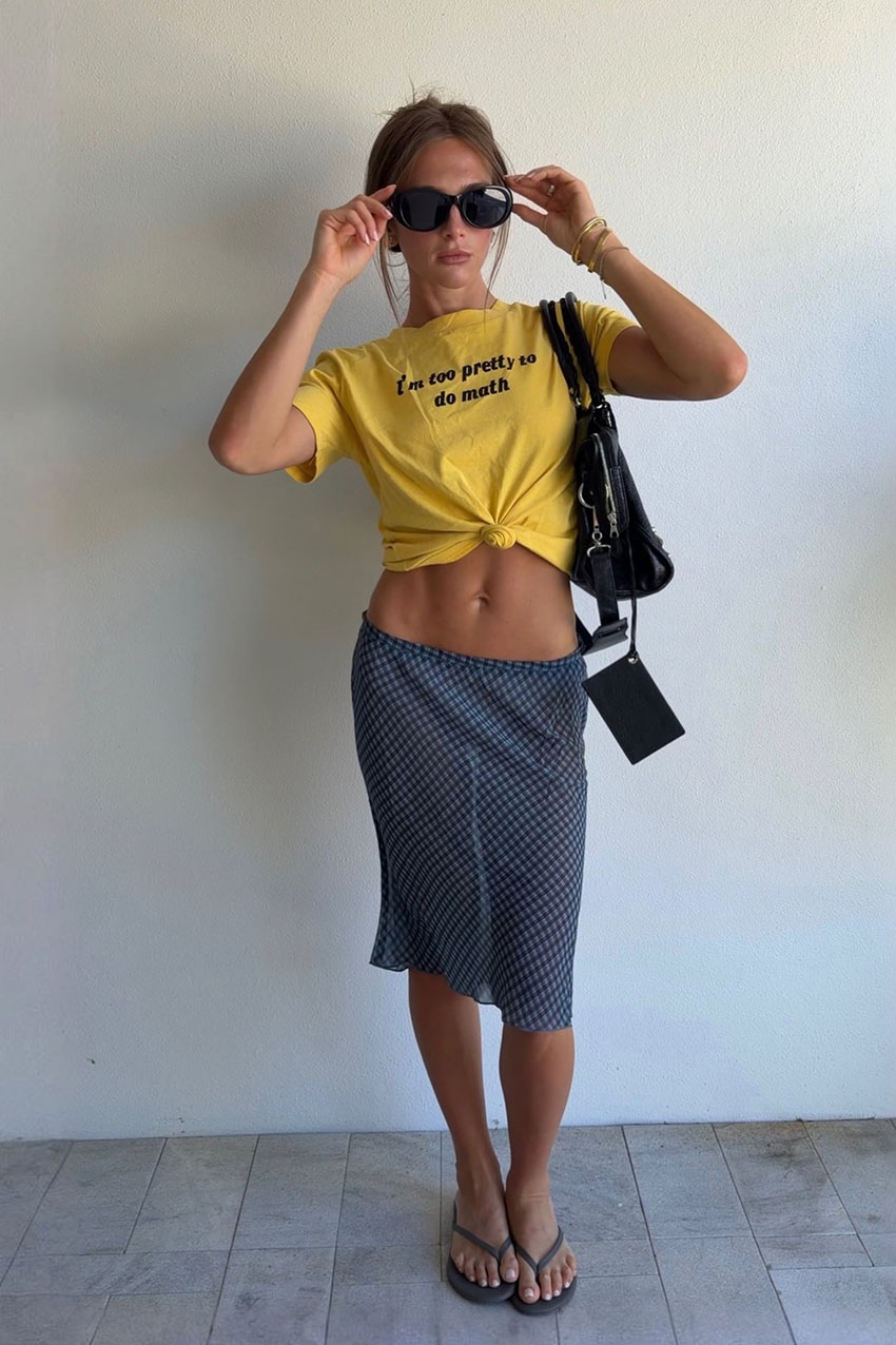 A woman wearing a yellow graphic t-shirt, a plaid sheer skirt, and black flip flops.