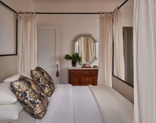 cream bedroom with white linen, canopy bed with beige curtains, and verdure print throw pillows with timber console in background with mirror and foliage in vase