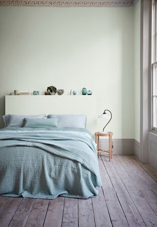Light green painted bedroom with sea green bedding, wood panel flooring and plants plus other trinkets on mantlepiece shelf space