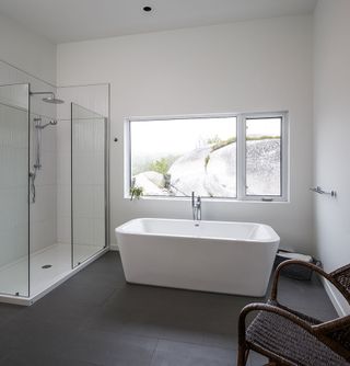 Interior view of the bathroom at Float House featuring white walls, grey tile flooring, a window, a white freestanding bath and a tiled shower with glass panels
