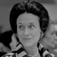 A black and white headshot of Wallis Simpson wearing a striped jacket and pearls looking to the left