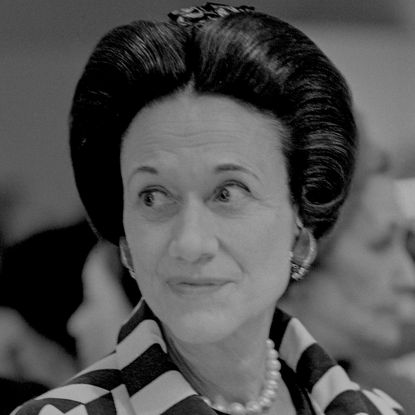 A black and white headshot of Wallis Simpson wearing a striped jacket and pearls looking to the left