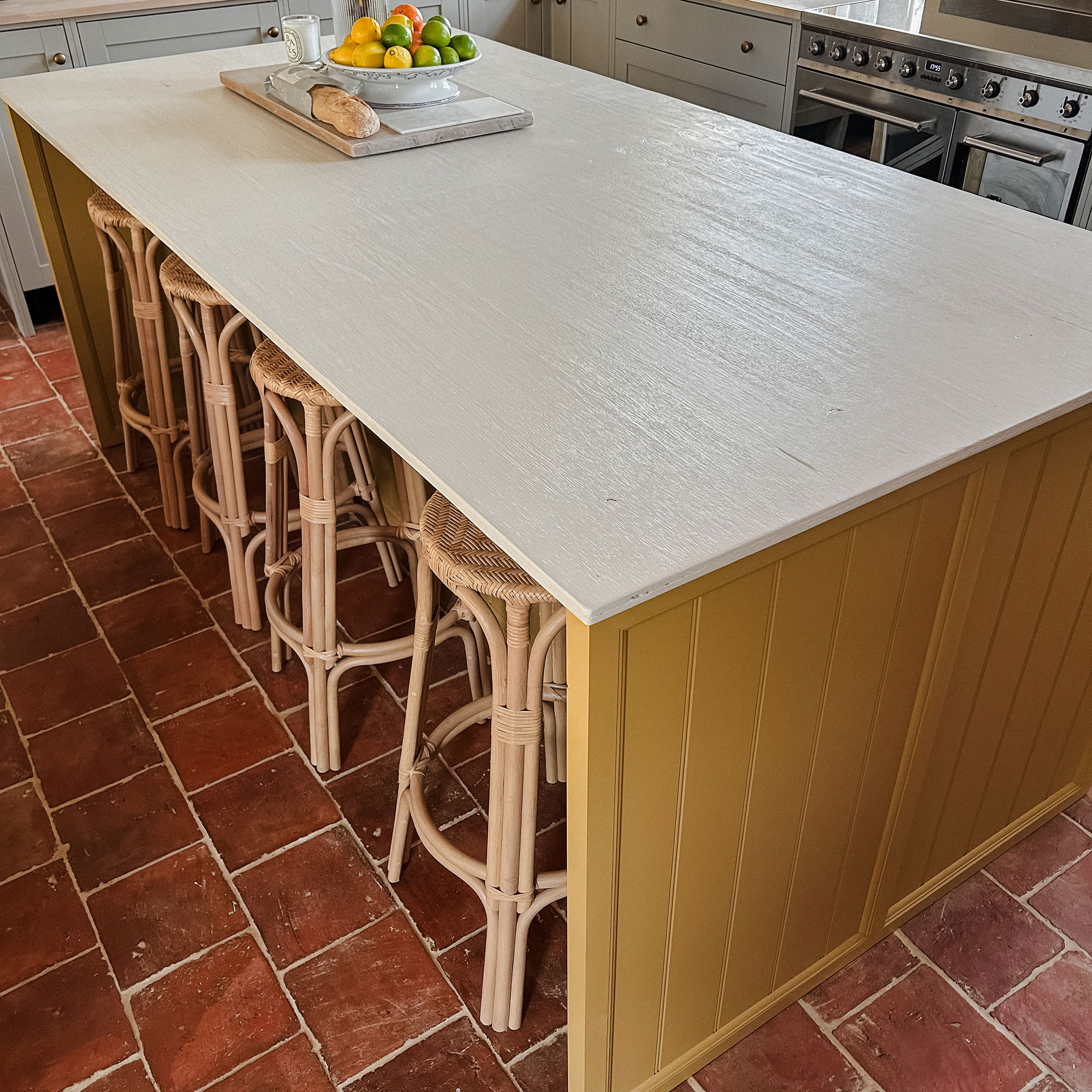 Kitchen island with white top and yellow patterned sides, with rattan bar stools, on tiled terracotta floor