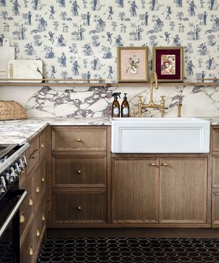 Victorian looking kitchen with black hex floor tiles, wooden cabinets, marble countertop, Butler sink, artwork, and blue printed wallpaper