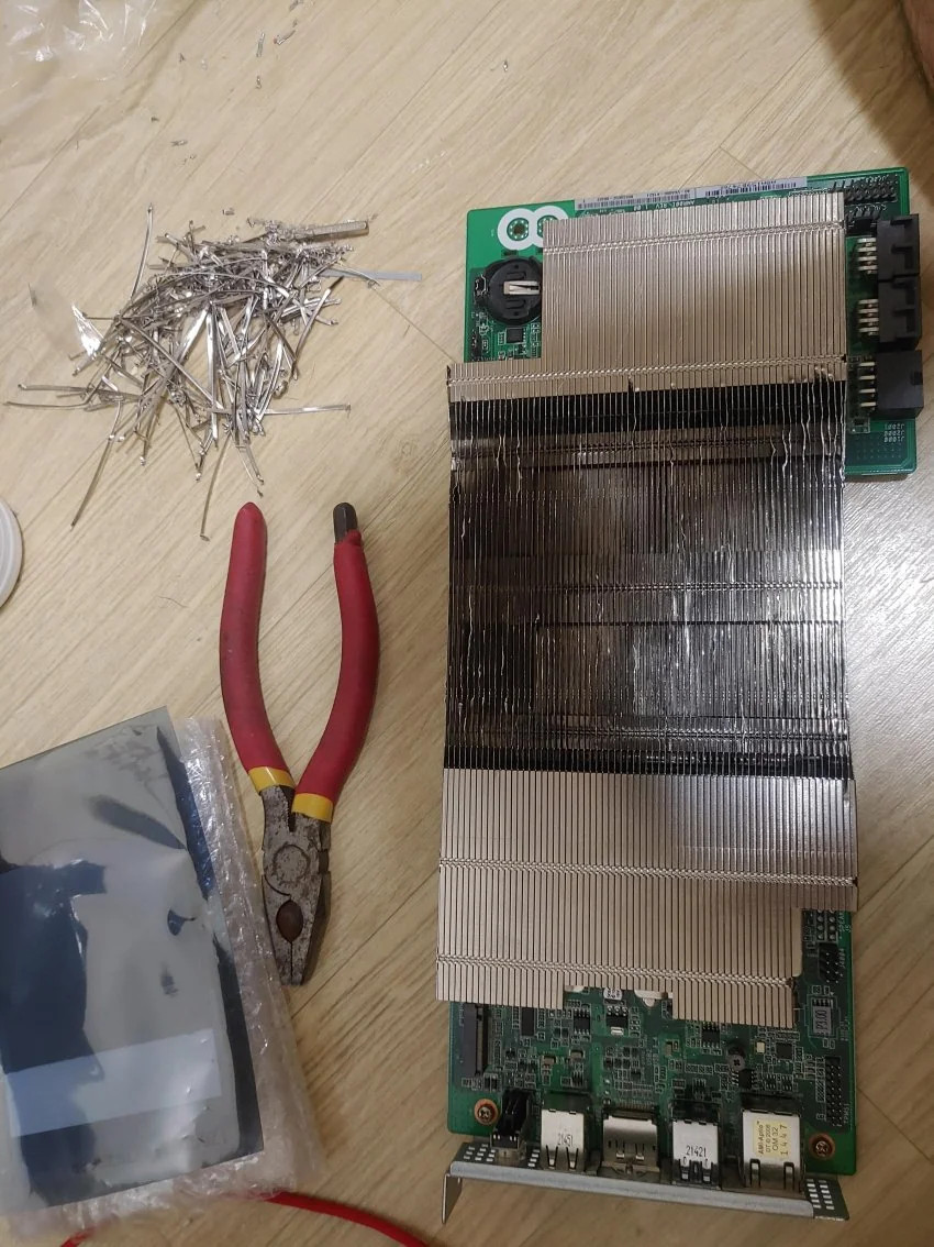 A BC250 computer circuit board with a large heatsink lies on a wooden floor. Nearby, a pair of red-handled pliers and a pile of metal shavings are visible.