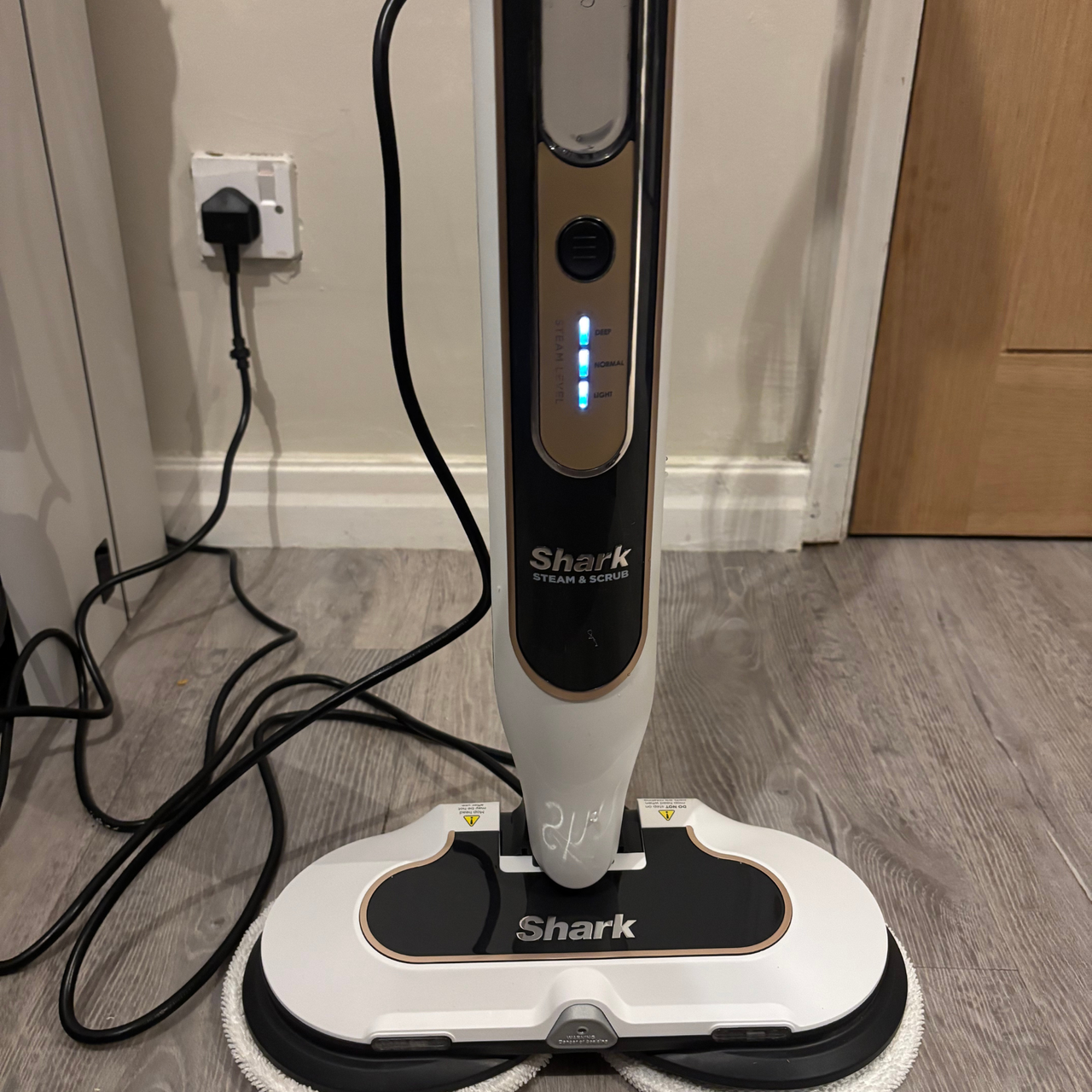 Shark Steam &amp; Scrub placed on hardwood floor in front of wall and plugged into mains