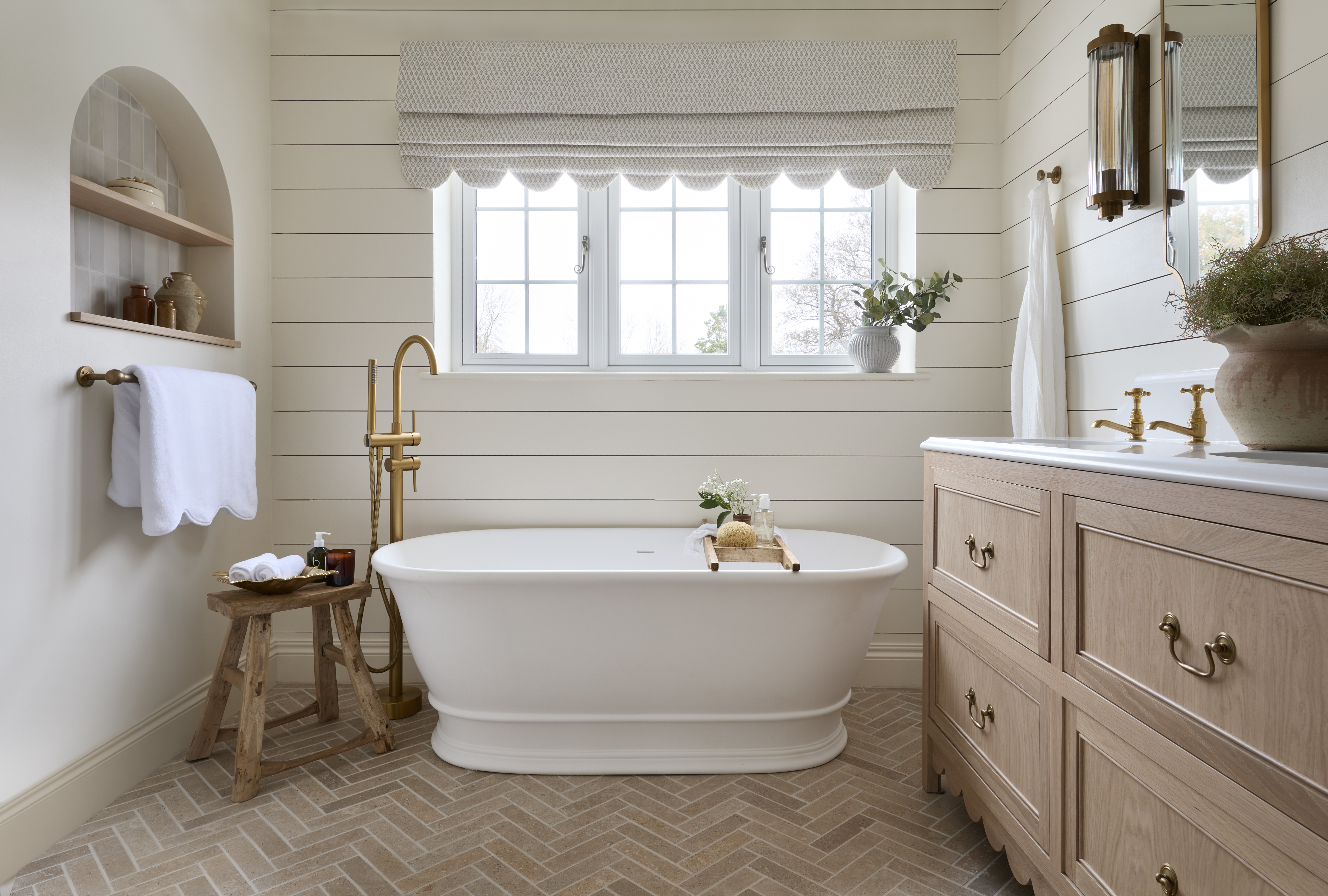 A guest bathroom with a large tub in the middle beneath a window. Beside the bath tub is a side chair and a towel rail with a towel on top. You can also see a corner view of the vanity and drawers.