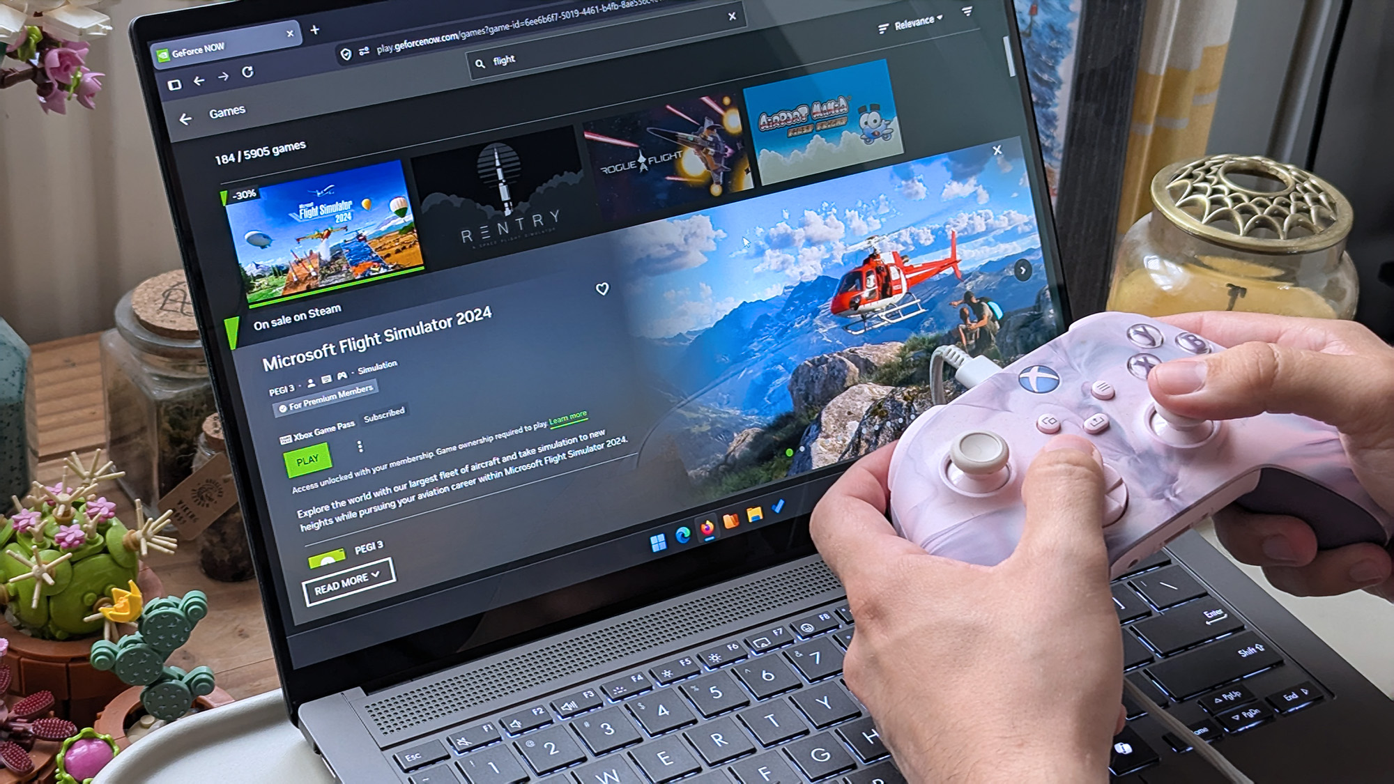 Hands holding a pink‑and‑white Xbox controller in front of an ASUS laptop displaying the GeForce NOW page for Microsoft Flight Simulator 2024, showing a green &ldquo;Play&rdquo; button.