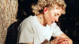 Blond Caucasian runner wearing Suunto Spark headphones checks her running watch as she leans against a tree