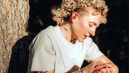 Blond Caucasian runner wearing Suunto Spark headphones checks her running watch as she leans against a tree
