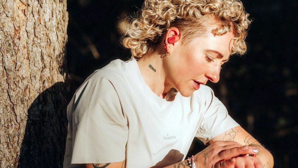 Blond Caucasian runner wearing Suunto Spark headphones checks her running watch as she leans against a tree