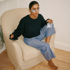 A woman sitting in a chair wearing under-eye skincare patches drinking a glass of water