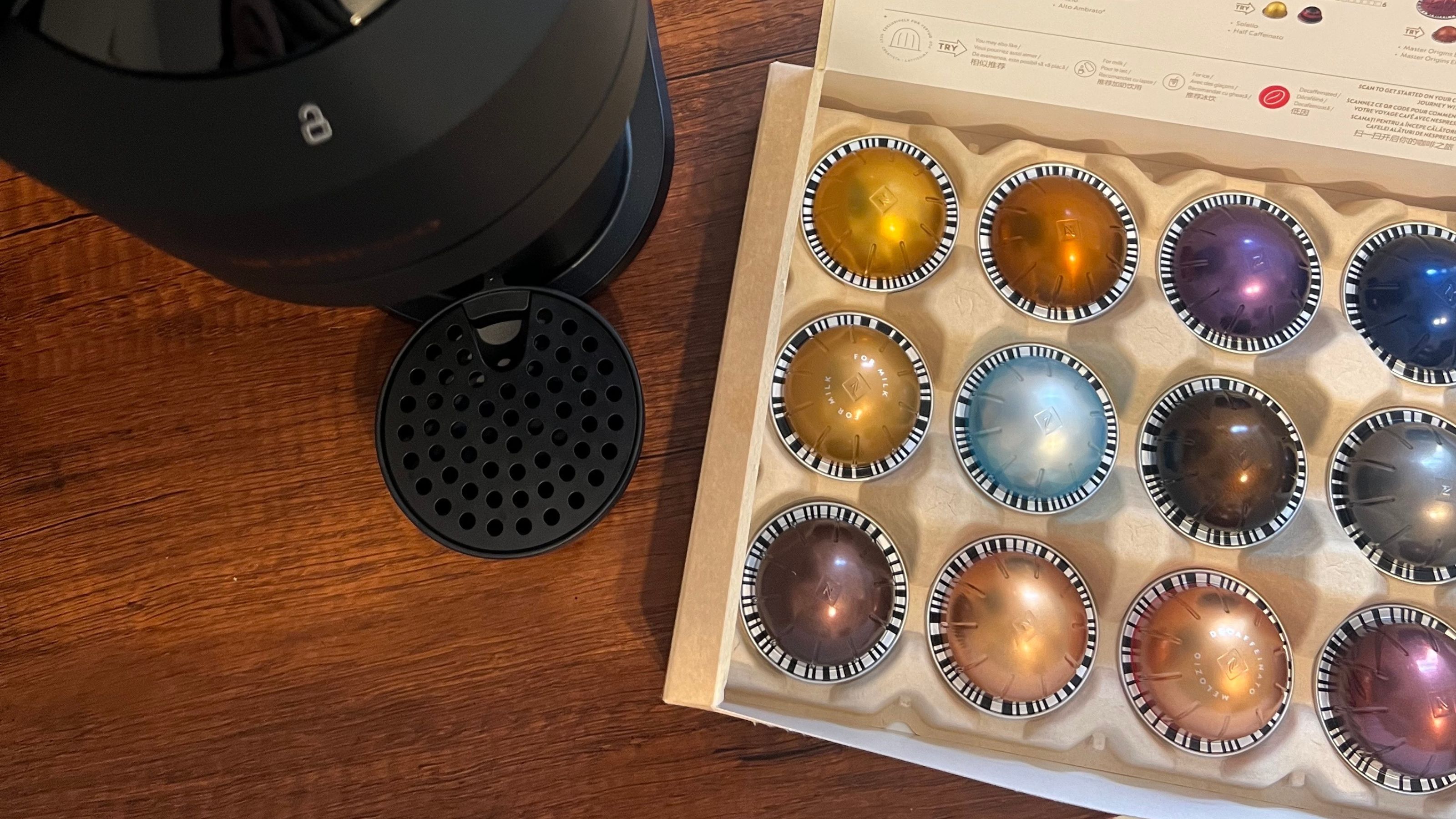 Close-up image of a black Nespresso machine on a wooden countertop with a box of assorted coffee pods next to it.