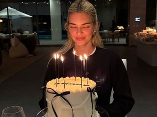 Kendall Jenner posing with her 29th birthday cake in a black cardigan and chunky gold earrings.