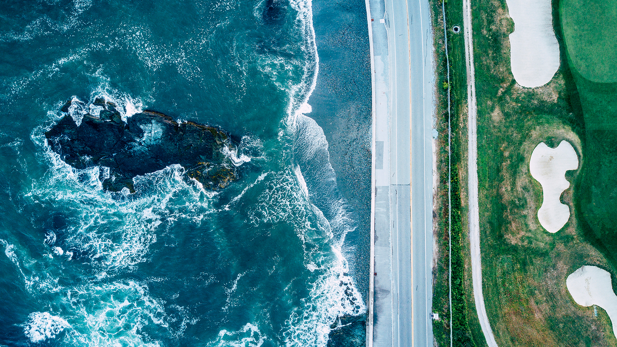 Aerial view of a coastal area with crashing waves on dark rocks to the left and a road with manicured golf course and sand bunkers to the right. Vibrant contrast