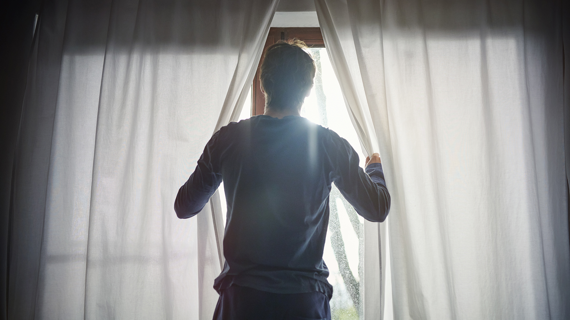 Un hombre abre las cortinas de las ventanas de su dormitorio y deja entrar la luz de la mañana.