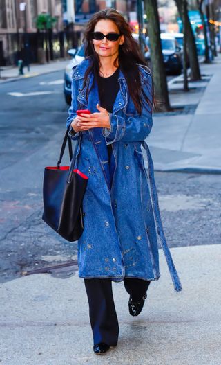 Katie Holmes walks down the street wearing a denim trench coat and studded flats