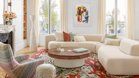 Living room with white curved corner sofa, round wood and white coffee table, red patterned rug and white accessories