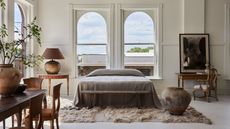 A bed with gray bedding and no headboard in front of double windows in a large, luxury white bedroom. There are antiques around the room and a shag rug.