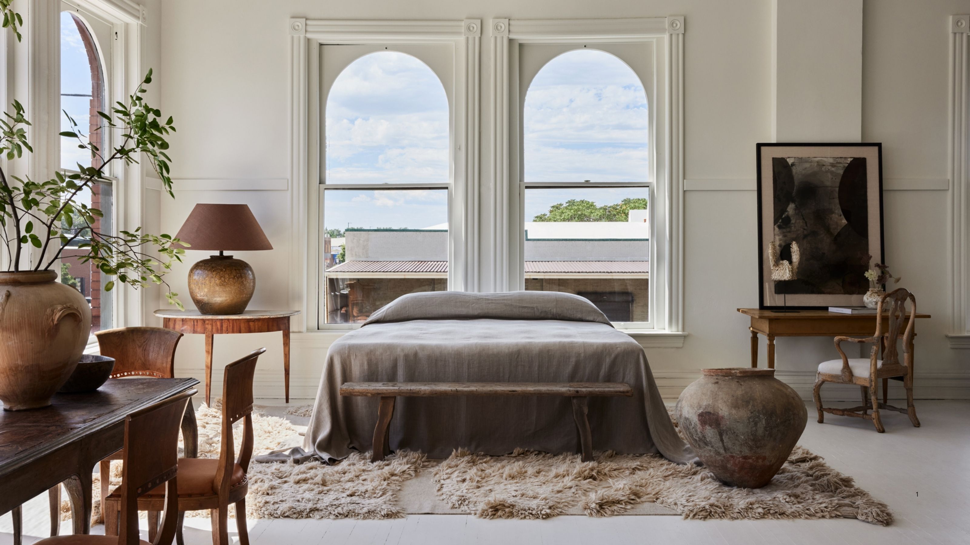 A bed with gray bedding and no headboard in front of double windows in a large, luxury white bedroom. There are antiques around the room and a shag rug.