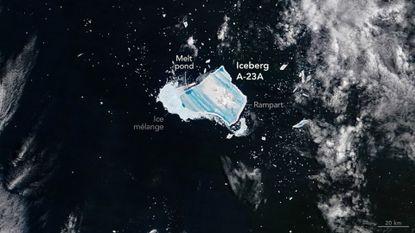 A blue jagged shape is seen in dark waters with white foam from the sea with the large white label Iceberg A-23A next to the blue shape