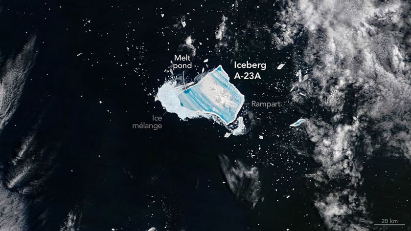 A blue jagged shape is seen in dark waters with white foam from the sea with the large white label Iceberg A-23A next to the blue shape