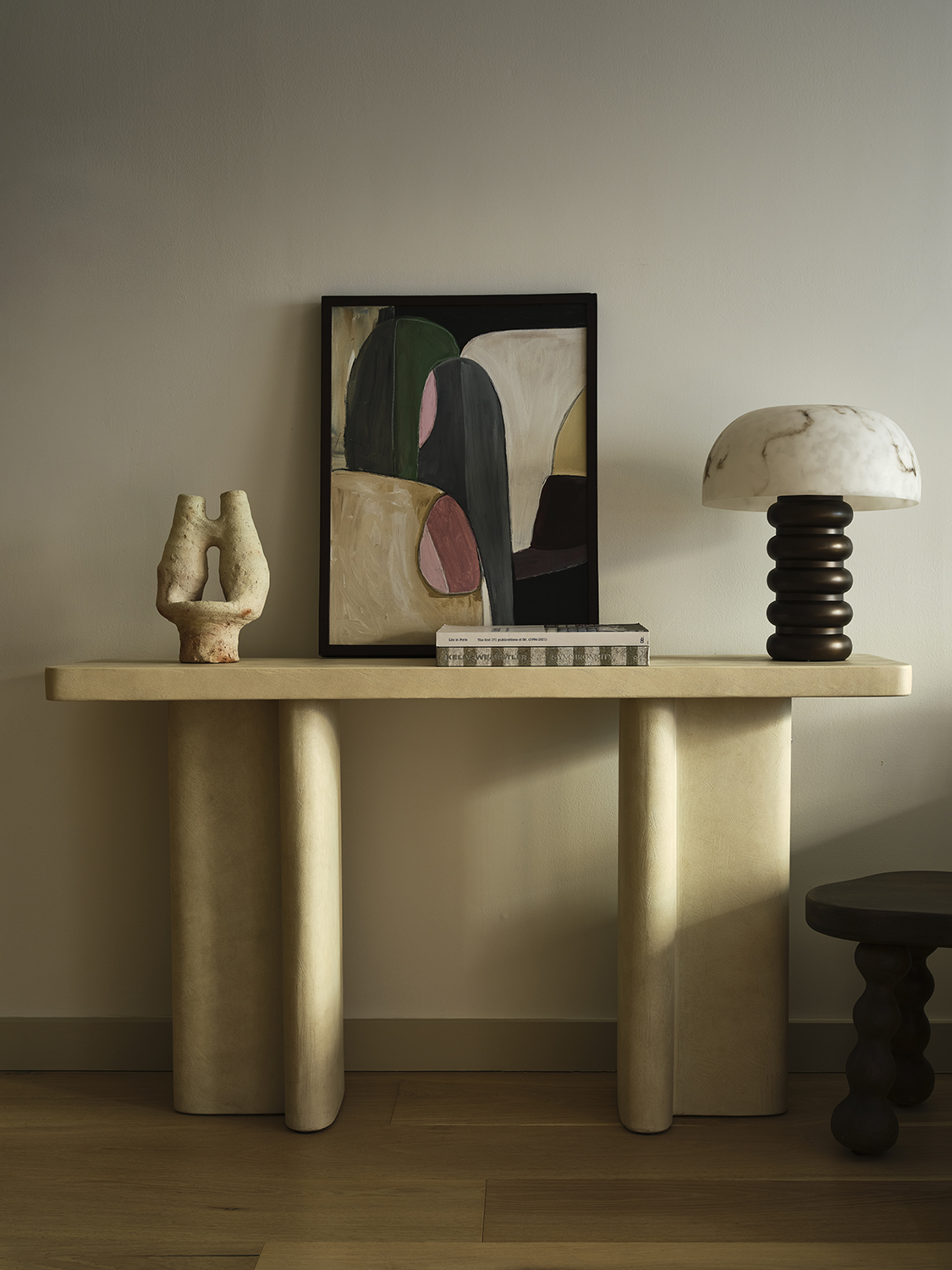 Image of a cream console table in a white entryway. There is an abstract art piece, a black and white lamp, magazines, and a sculptural object on the table.