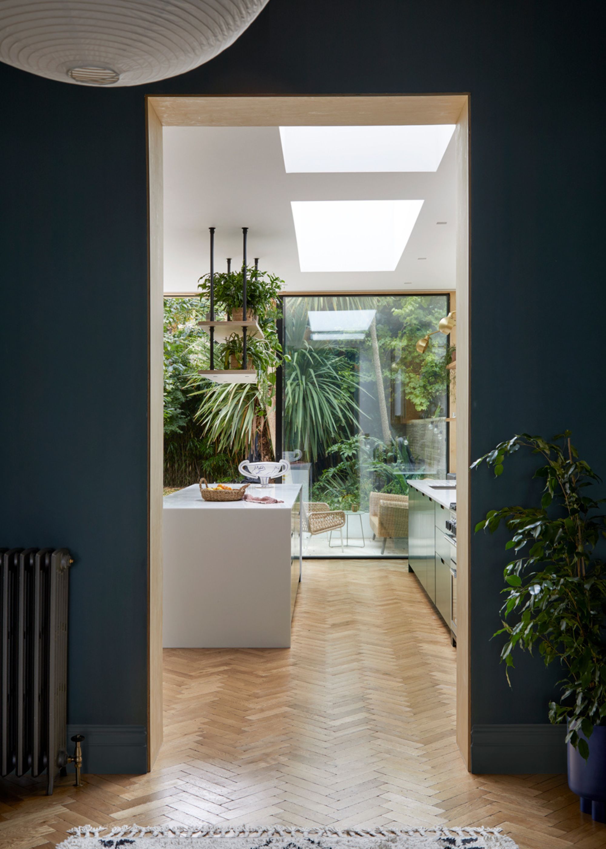 An entrance leading to the kitchen with the view of the garden from floor to ceiling glass doors. There is also a large island in the kitchen with open shelving and plants hanging from it.