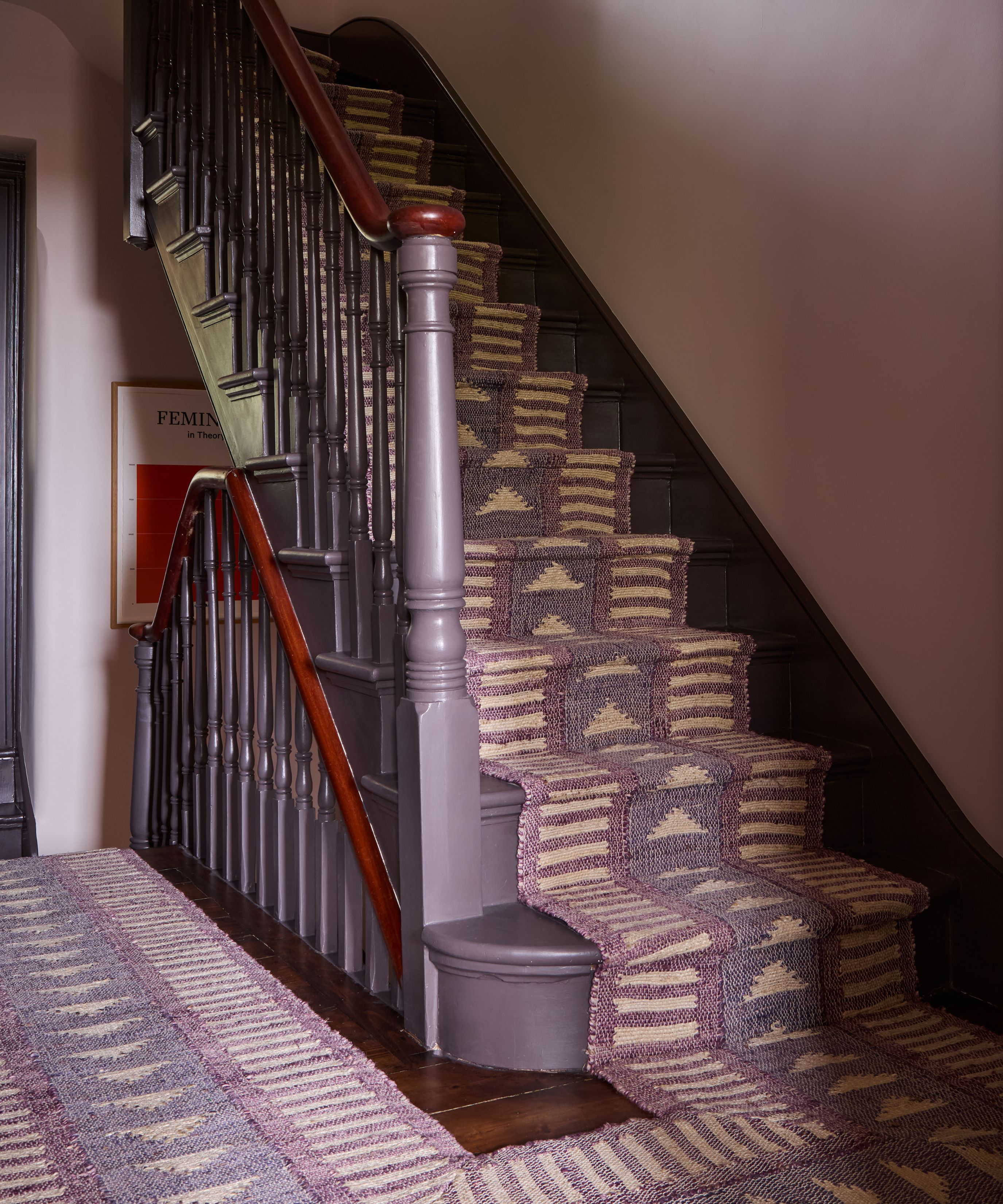 A classic wooden staircase painted in a deep mauve-purple, featuring a decorative woven runner with cream and purple geometric patterns. A matching rug extends across the dark wood floor at the base of the stairs.
