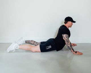 Coach Corey Perkins performs a Cobra pose. He is lying on the floor, with his legs behind him and his torso propped up, held up by his arms. He wears shorts, a t-shirt, a black baseball cap and sneakers.