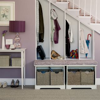 lilac walls and coat cubbies in under stairs storage area, with bench seat in front and wicker baskets underneath