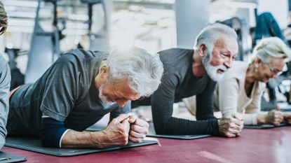 Three seniors in a gym in a row performing the plank exercise. One man is resting on the floor, unable to continue