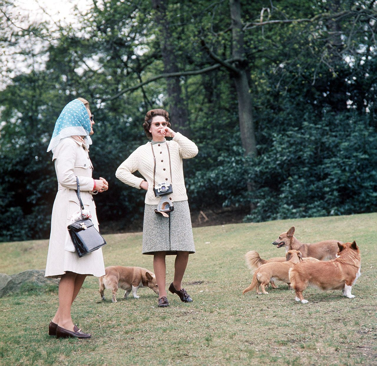 Queen Elizabeth II stands outdoors with another woman, surrounded by several corgis on a grassy lawn.