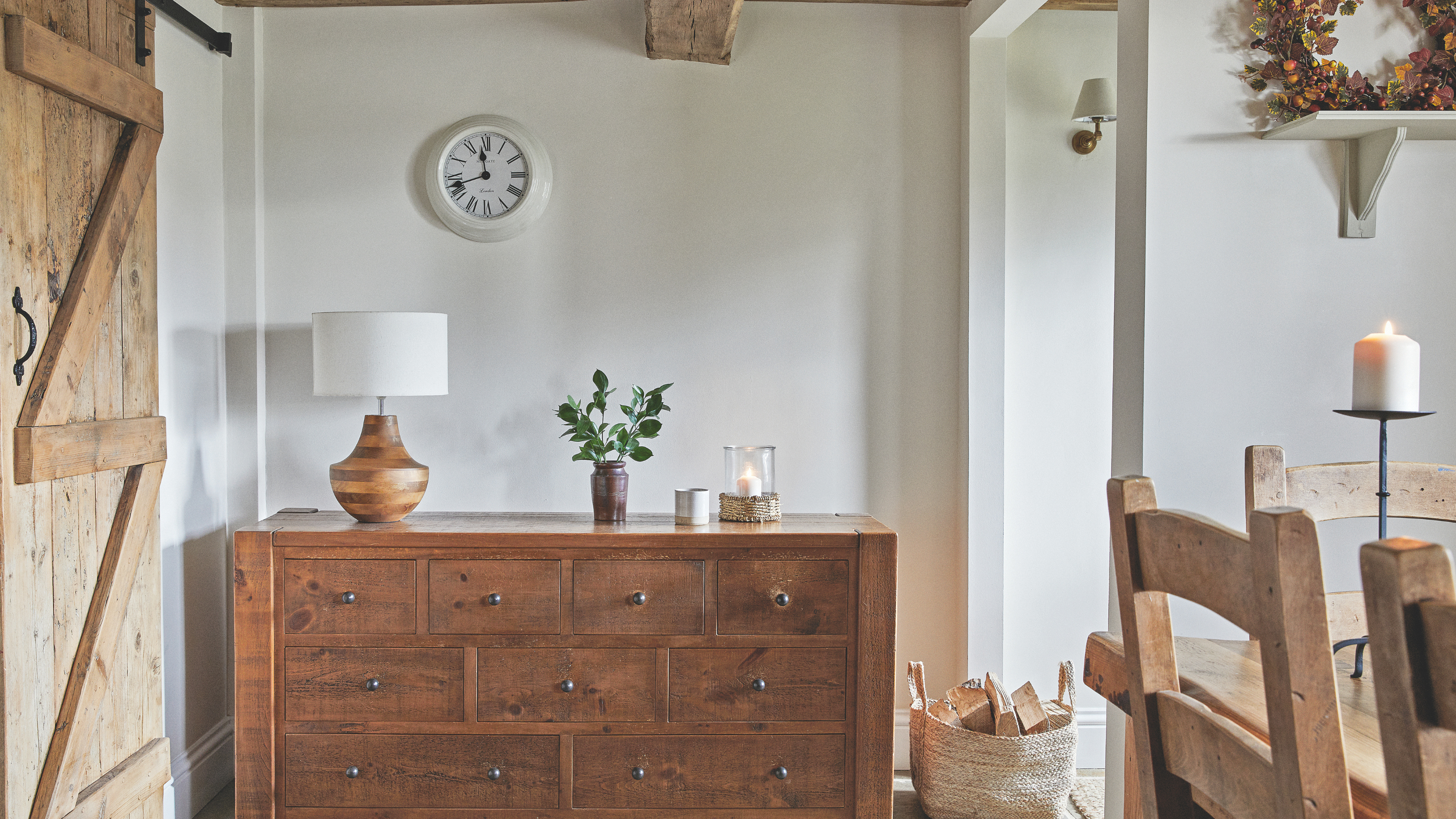 Two lit candles on a wood side board. There is a small potted plant and wood and white fabric lamp.