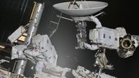 two astronauts in bulky white space suits maneuver on the outside of a large facility covered in solar panels and machinery