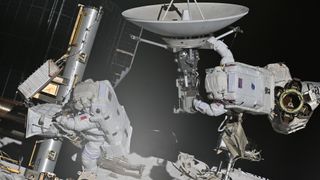 two astronauts in bulky white space suits maneuver on the outside of a large facility covered in solar panels and machinery