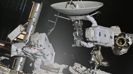 two astronauts in bulky white space suits maneuver on the outside of a large facility covered in solar panels and machinery