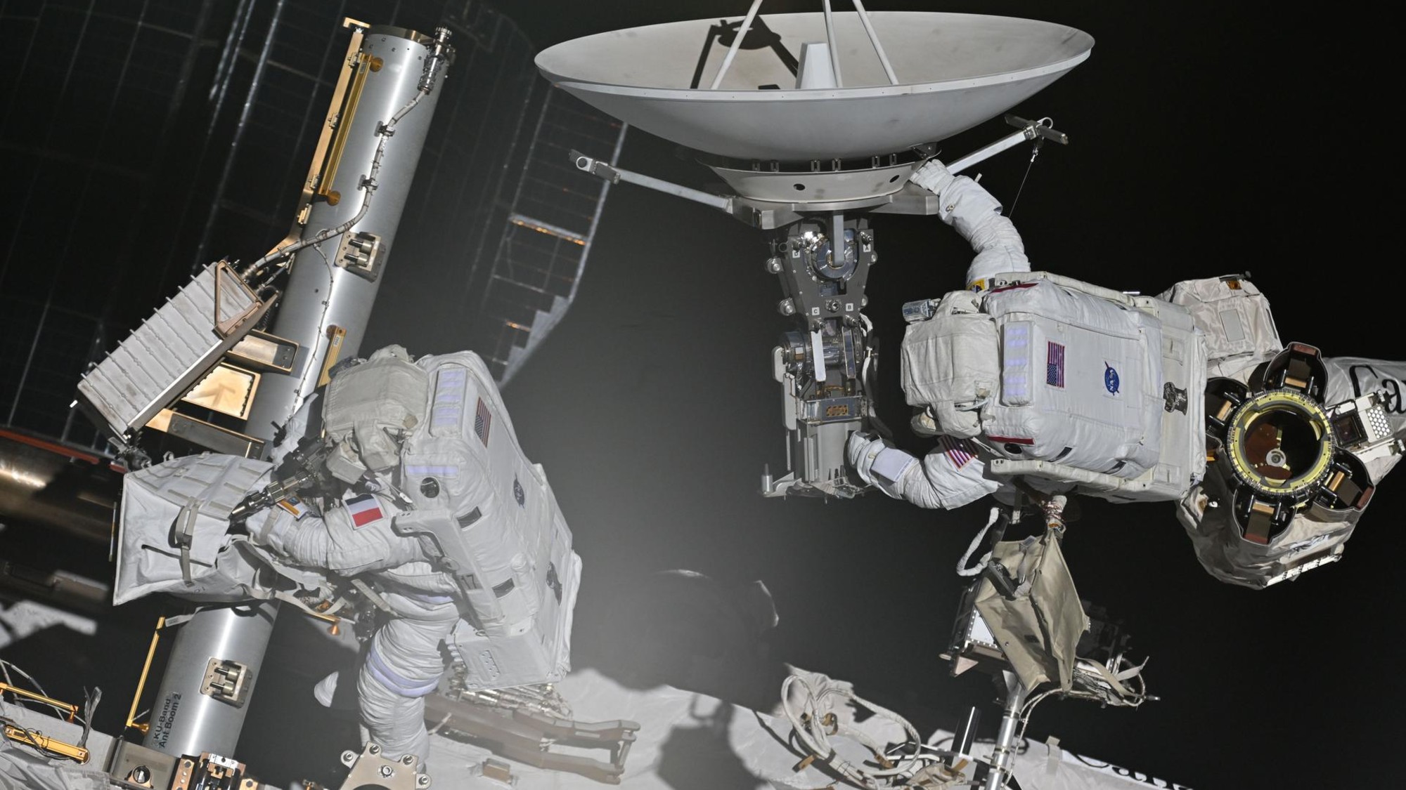 two astronauts in bulky white space suits maneuver on the outside of a large facility covered in solar panels and machinery
