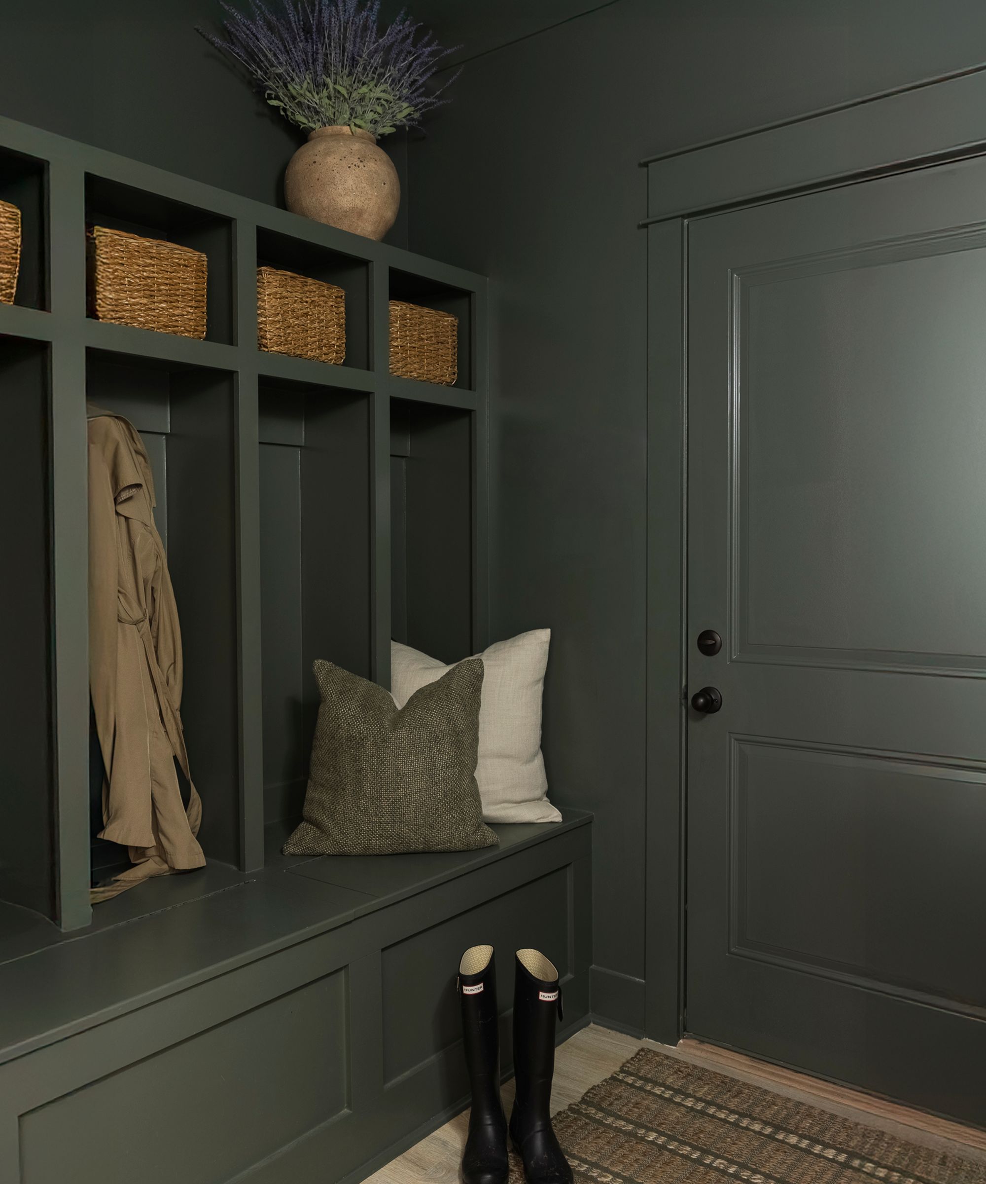 Mudroom painted in Sherwin-Williams Pewter green, with a built-in bench and storage, rattan baskets, a jute rug, and a pair of Hunter Wellingtons.