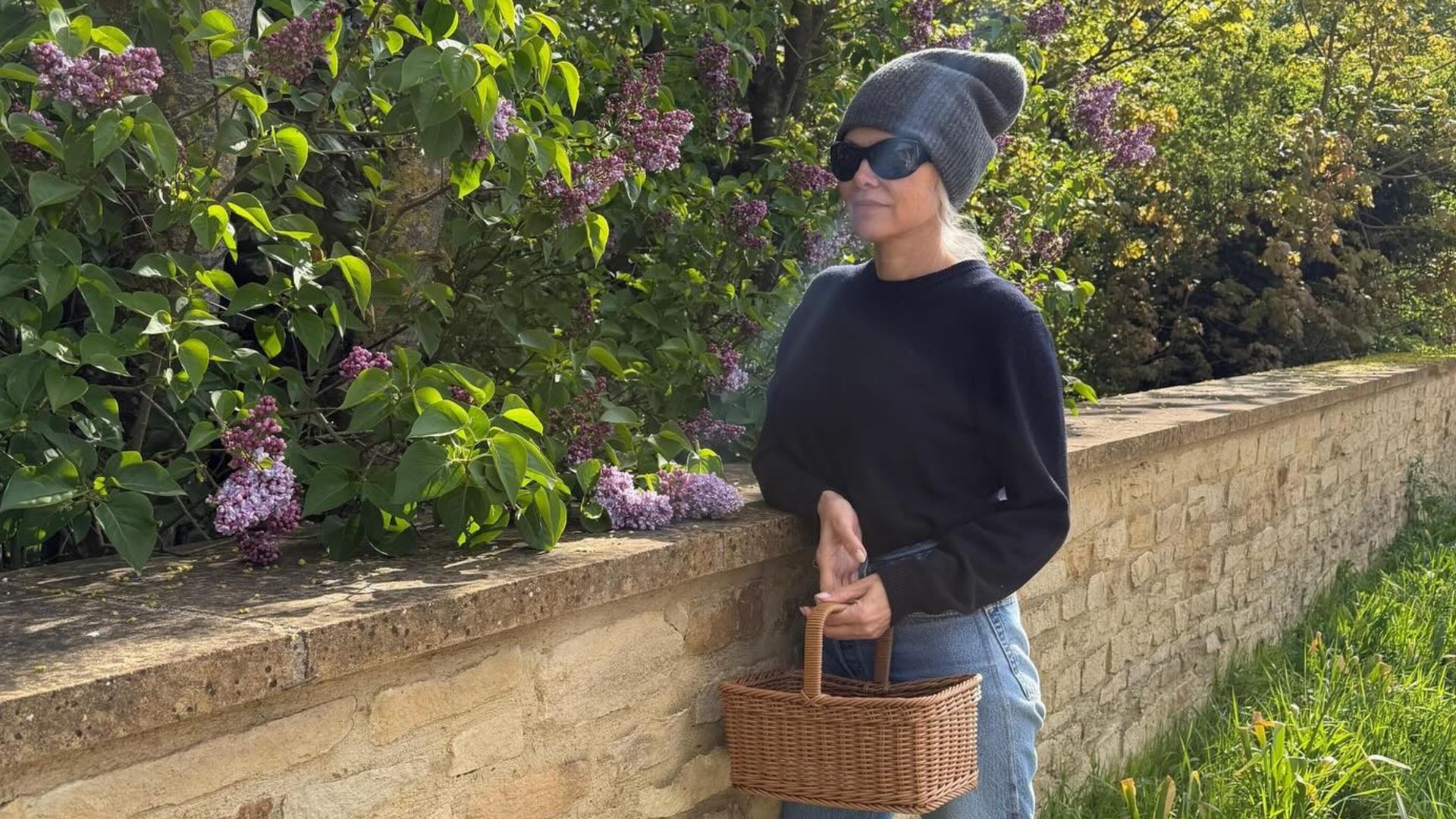 Image of Pamela Anderson in a garden wearing jeans with a navy jumper, beaning and black sunglasses.