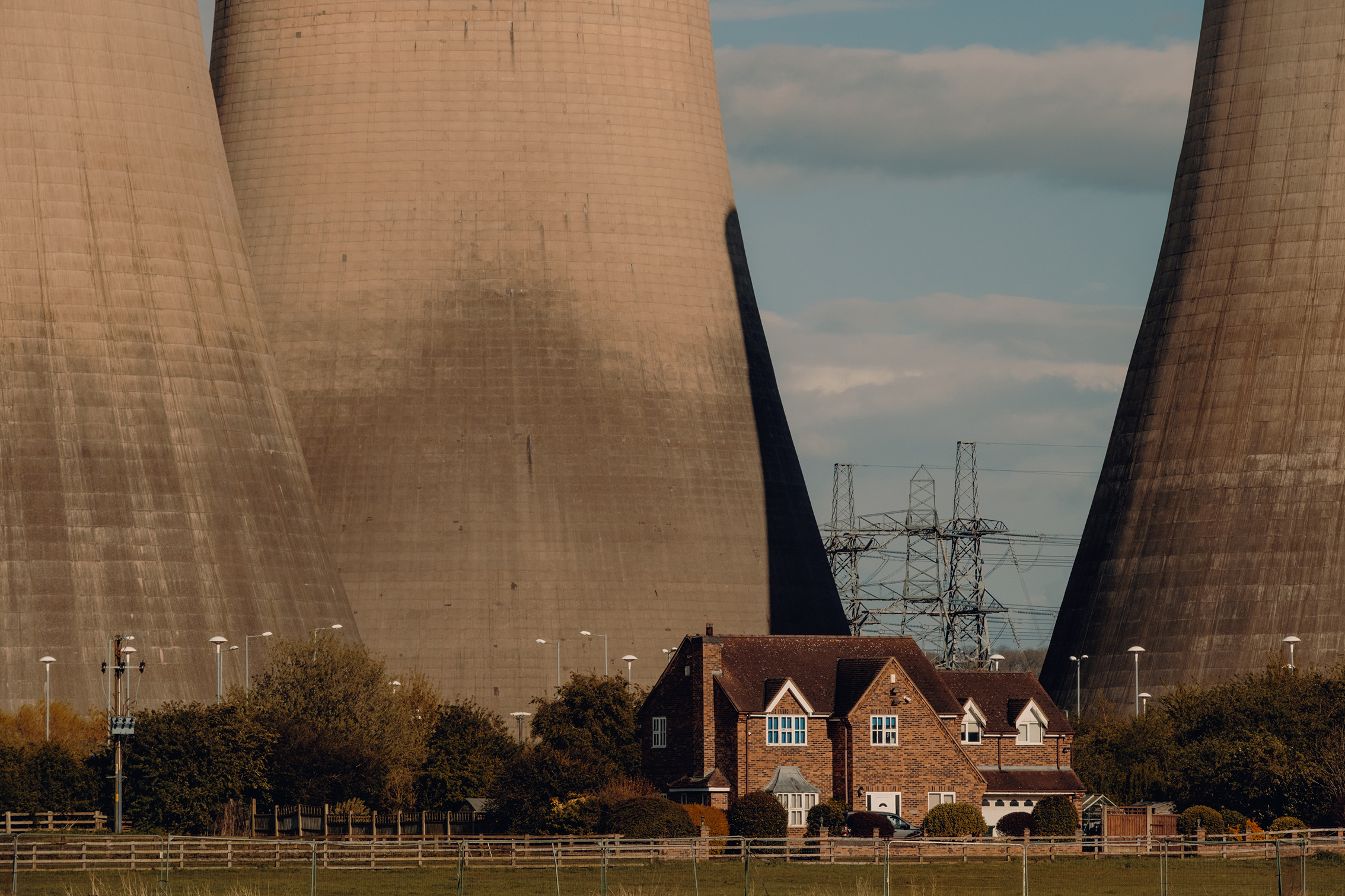 A small house surrounded by trees, nestled between two large cooling towers and power lines against a cloudy sky