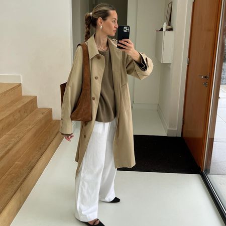 Fashion influencer Anouk Yve taking a mirror selfie in a classic fall outfit, wearing a relaxed trench coat, brown sweater, and white pants, black flats, and carrying a suede tote bag.