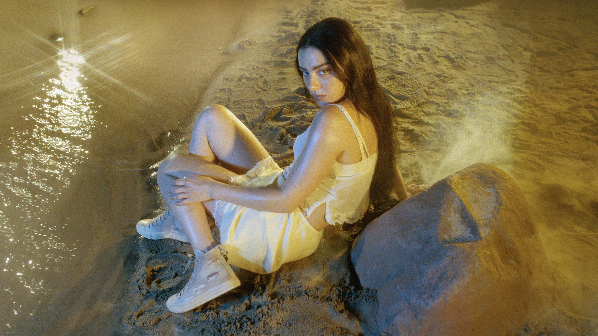 charli xcx lays in the sand in a white clothing set wearing bridal rose chuck taylor allstar
