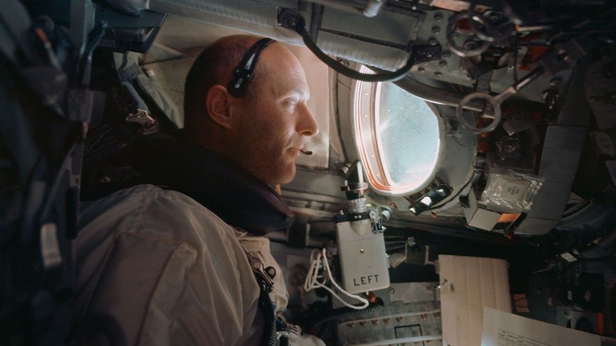 a man in a white flight suit inside a cramped spacecraft capsule filled with screens and wires