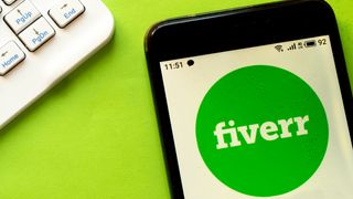 The Fiverr logo (the word &quot;Fiverr&quot; in white, lower case serif font on a green circle) shown on a phone sitting on a green surface. In the top left corner, the corner of a white computer keyboard can be seen, with only the arrow keys visible.