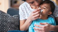 A mother with dark hair leans over a small dark-haired child wearing a bright blue shirt. The mother presses a white handkerchief to the child's nose.