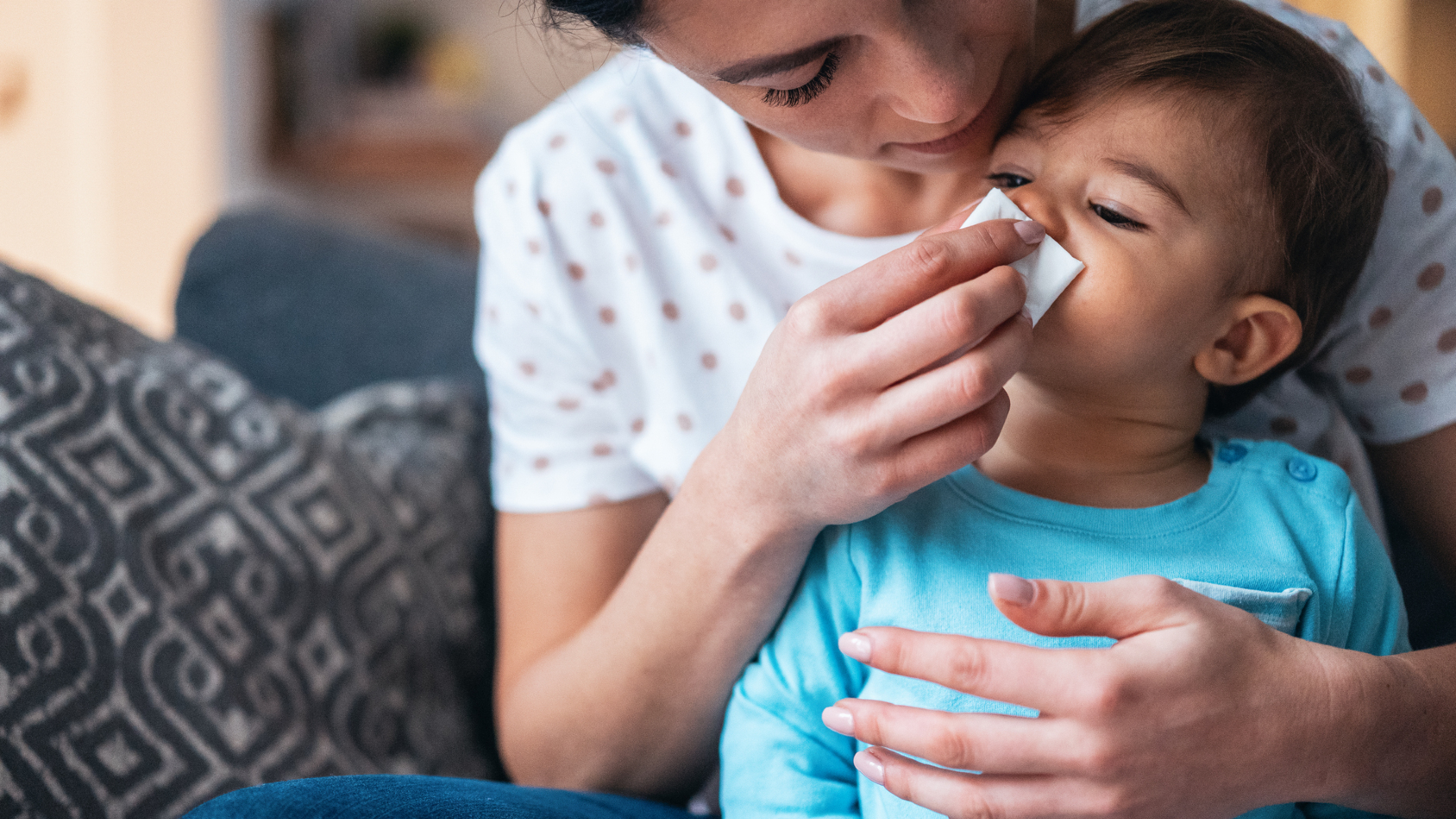 A mother with dark hair leans over a small dark-haired child wearing a bright blue shirt. The mother presses a white handkerchief to the child's nose.