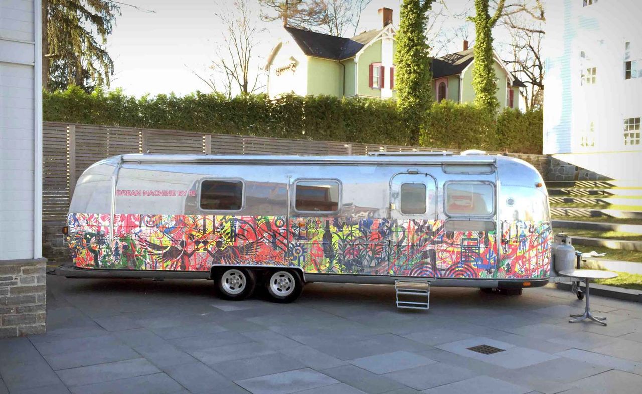 A campervan with graffitti designs on the body parked on an outside space on tiled concrete with houses in the background. Photographed during the day